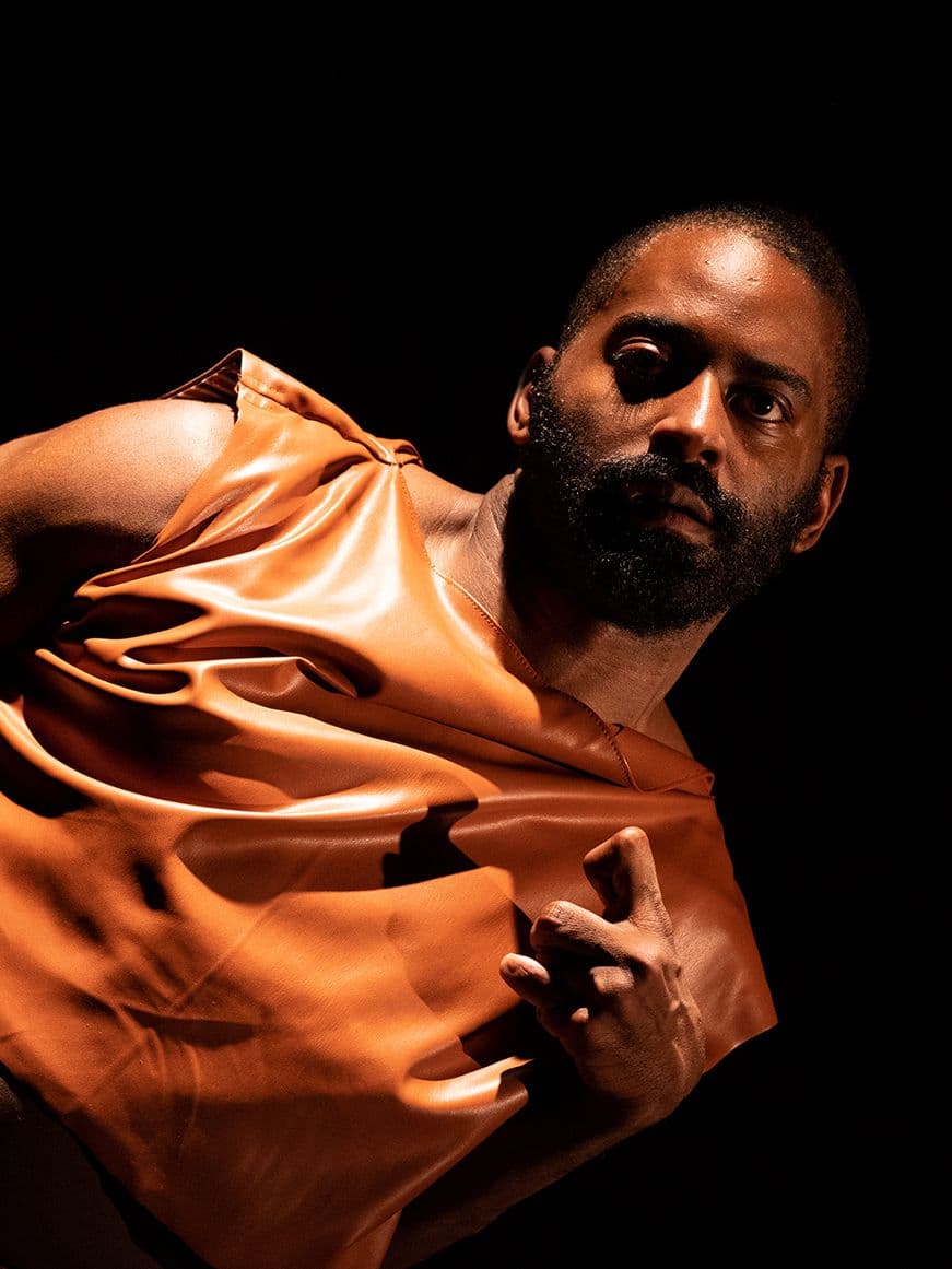 Jerron, a dark-skinned Black man leans with arm outstretched in a dark orange tunic By Maria Baranova